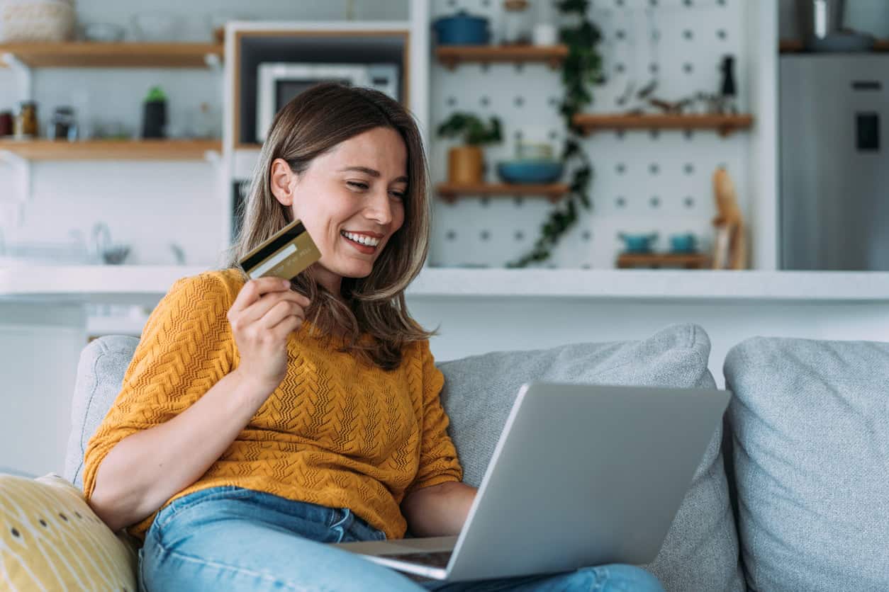 Woman smiling while using laptop and holding credit card, reading good news about a gold credit card offer at home