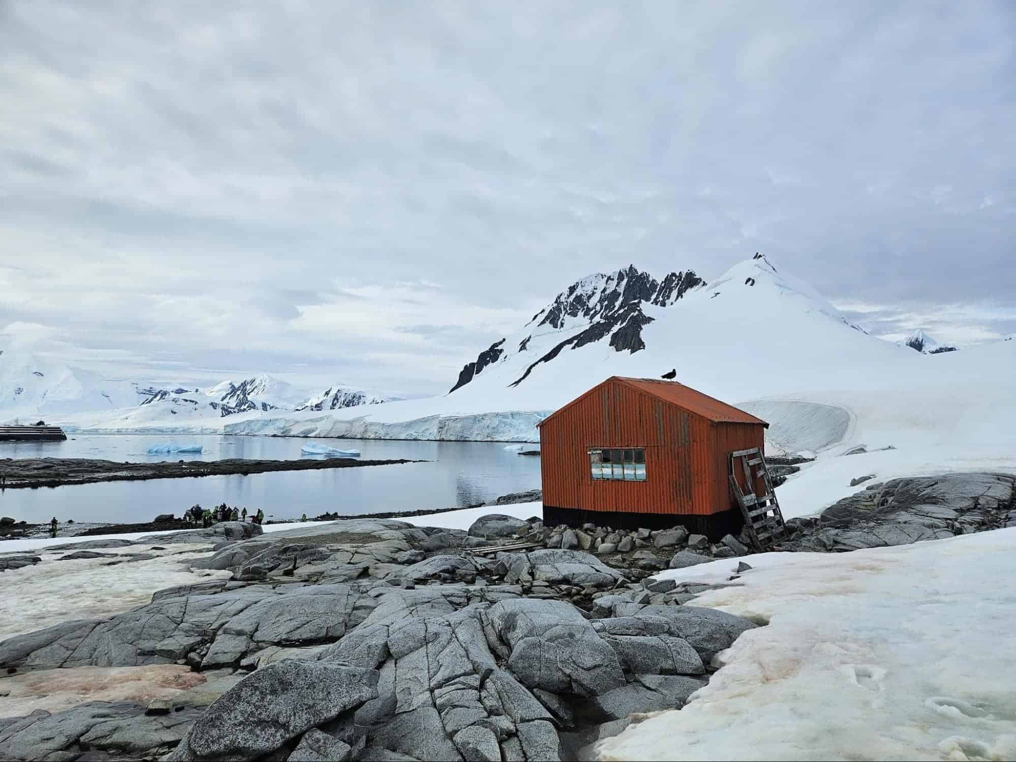 Small red expedition hut surrounded by snow-covered mountains, rocky terrain, and icy Antarctic waters during an Antarctica expedition | Photo by Anya Kartashova