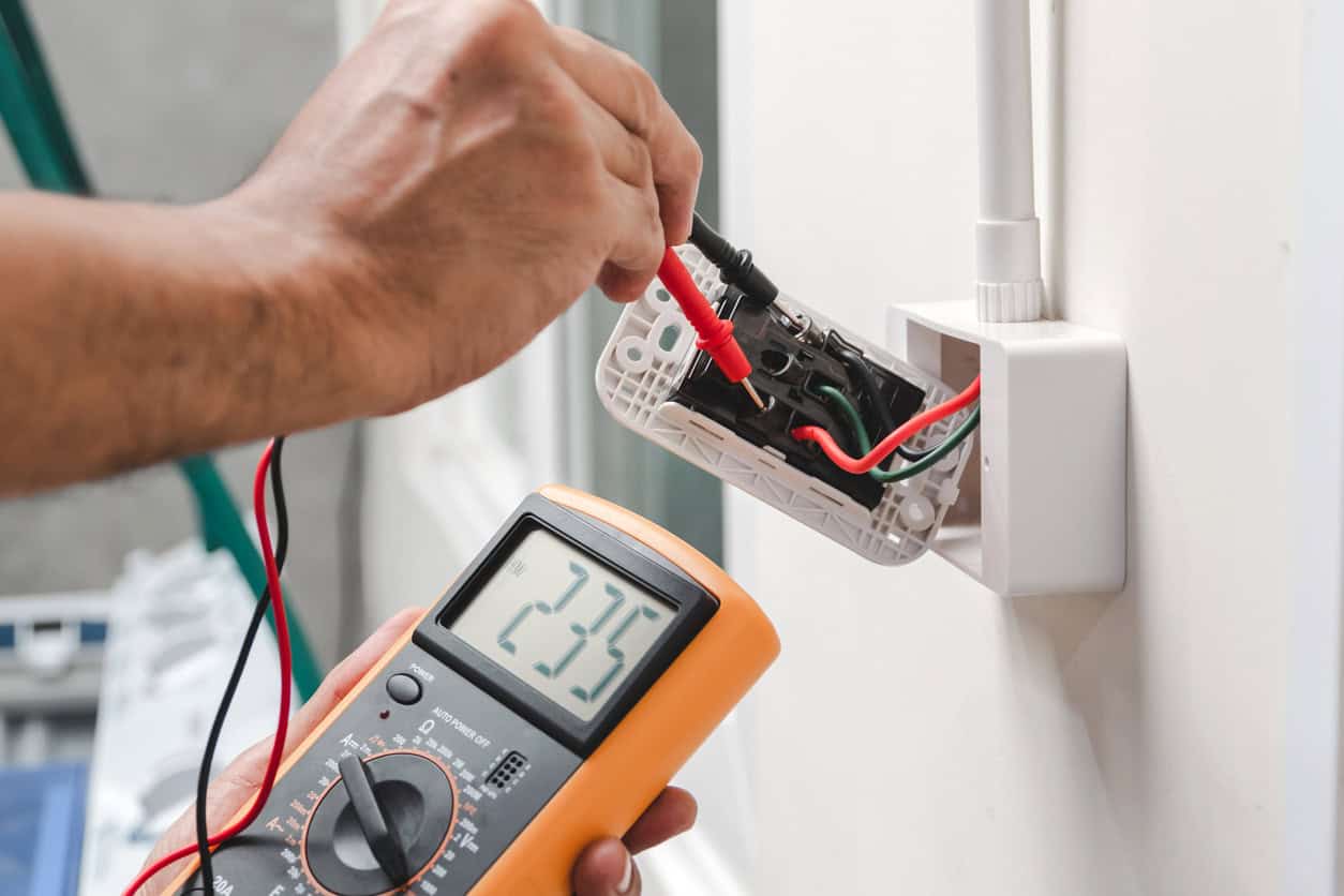 Electrician using a digital multimeter to test a wall outlet during an electrical installation.