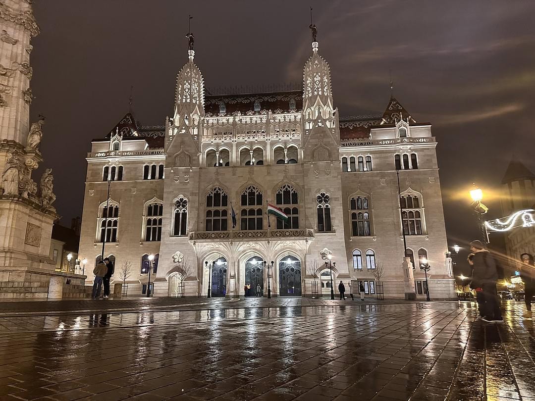 The illuminated Hungarian Parliament Building reflected on rain-soaked streets in Budapest at night, showcasing its striking Gothic Revival architecture.
