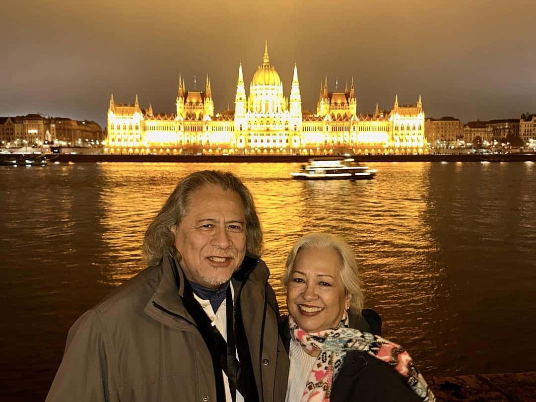 Travelers posing along the Danube River with the illuminated Hungarian Parliament Building glowing in the background during a night visit to Budapest.