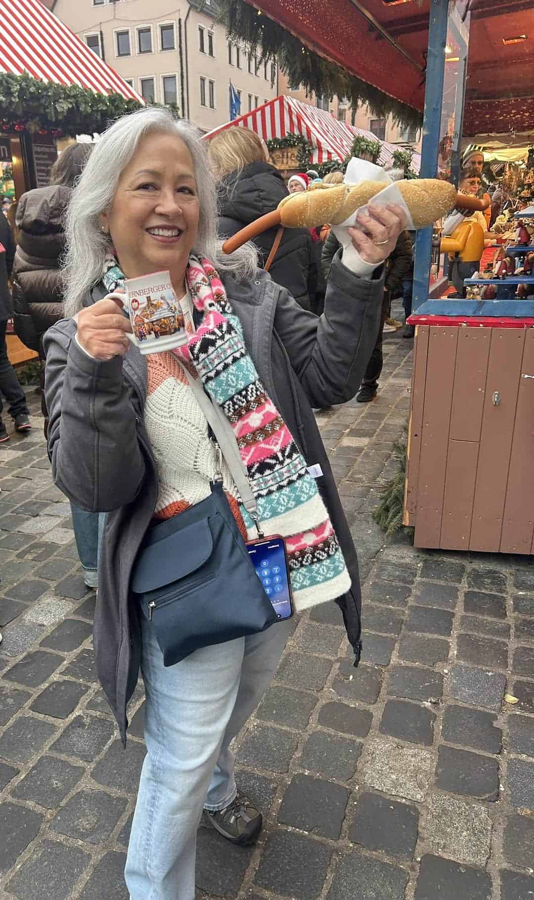Traveler holding a traditional Nuremberg bratwurst in a bread roll and a festive Christmas market mug while exploring the Nuremberg Christmas Market in Germany.