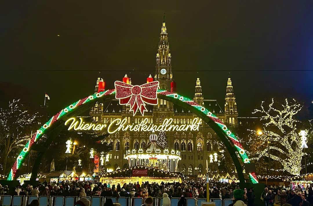 Night view of the Vienna Christmas Dream at Rathausplatz, featuring the illuminated Christkindlmarkt entrance, festive lights, carousel, and Vienna City Hall in the background.
