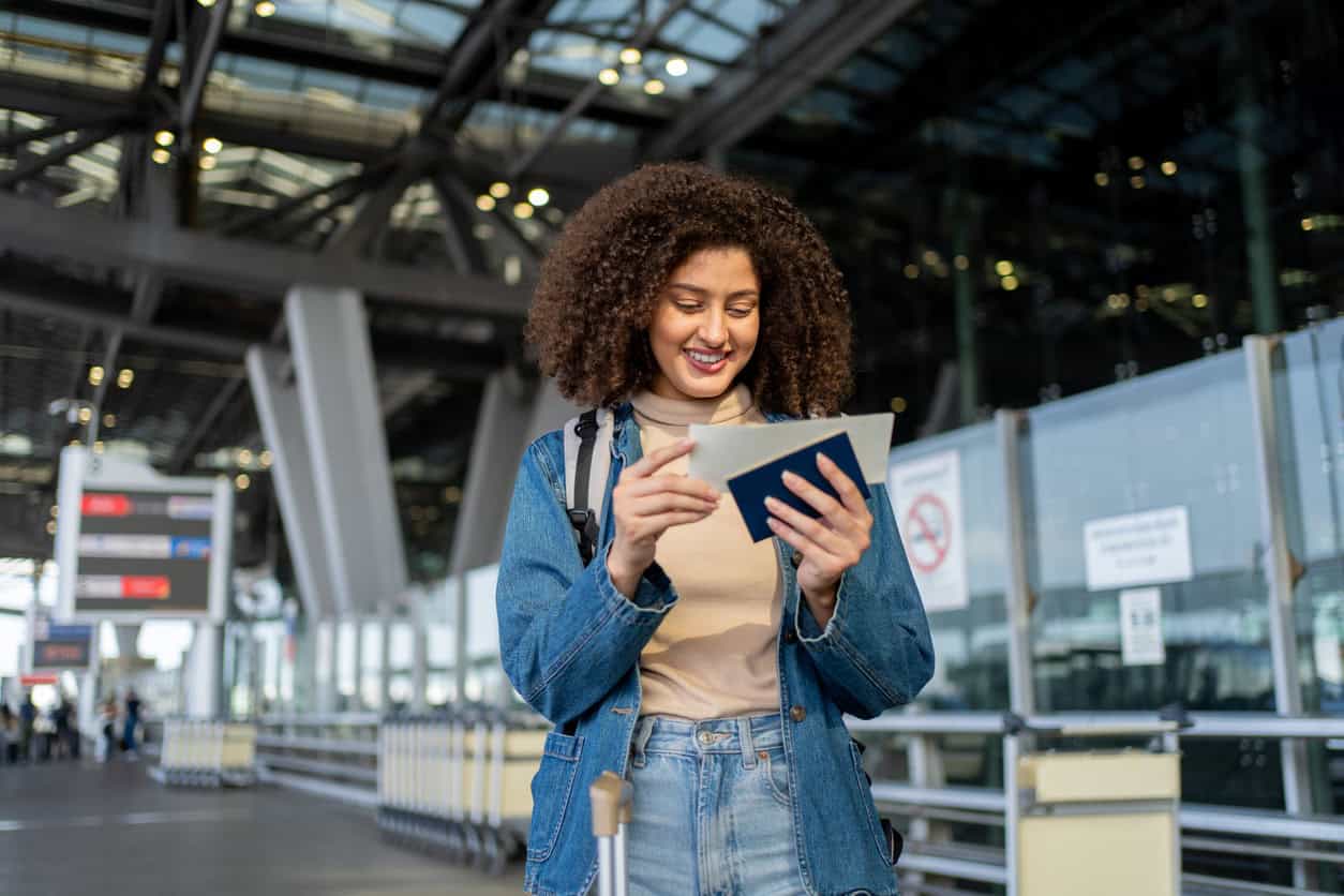 A traveler stands inside a modern airport terminal smiling while checking a passport and boarding pass before departure.