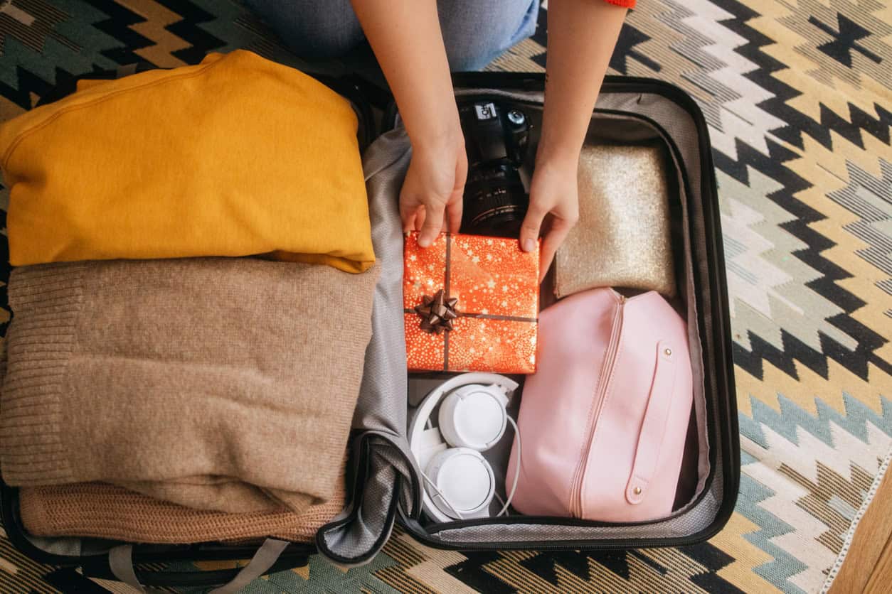 Open suitcase packed with clothes, headphones, a camera, toiletry bag, and a wrapped gift.