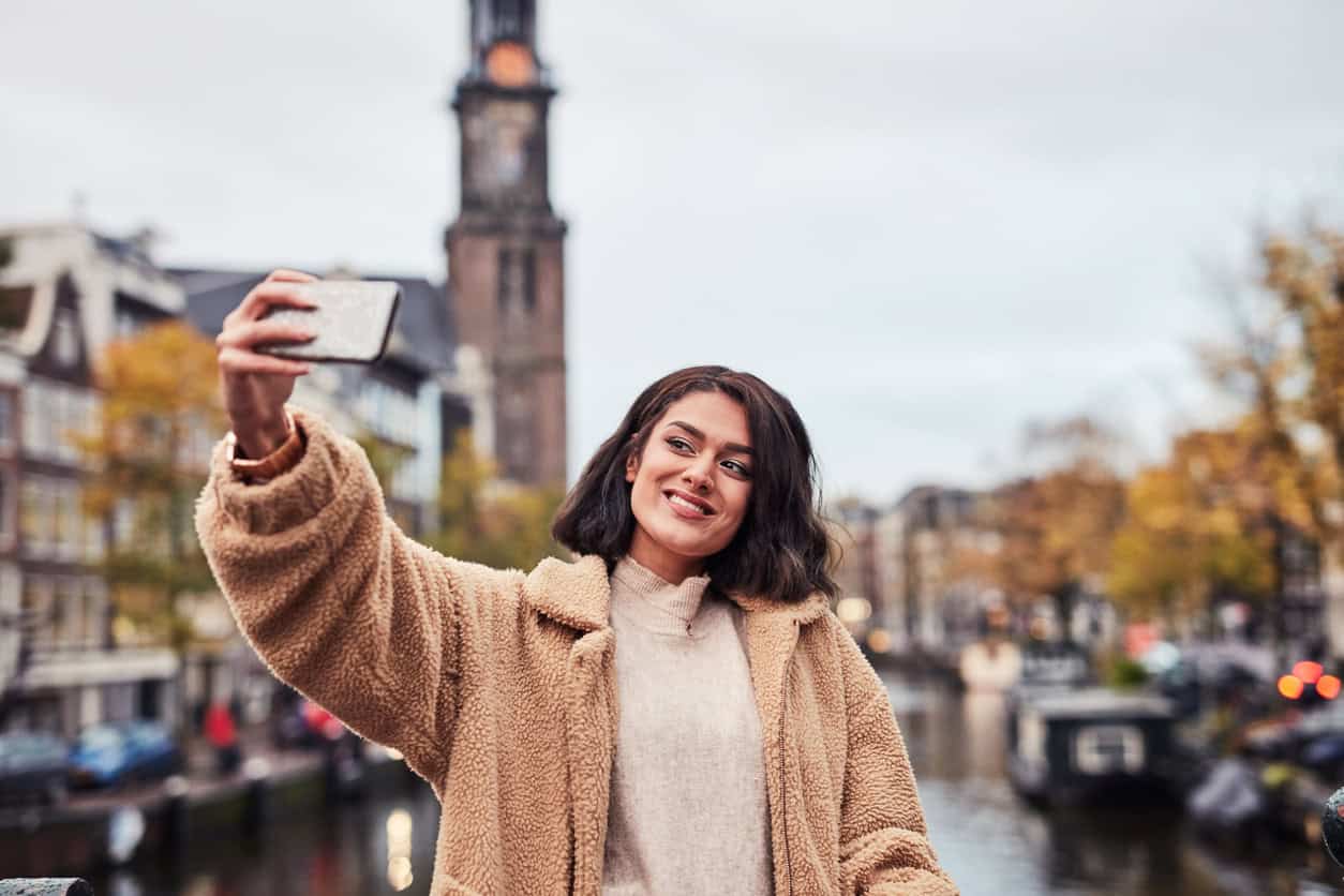 Woman taking a selfie while traveling in a European city with historic architecture and a canal in the background.